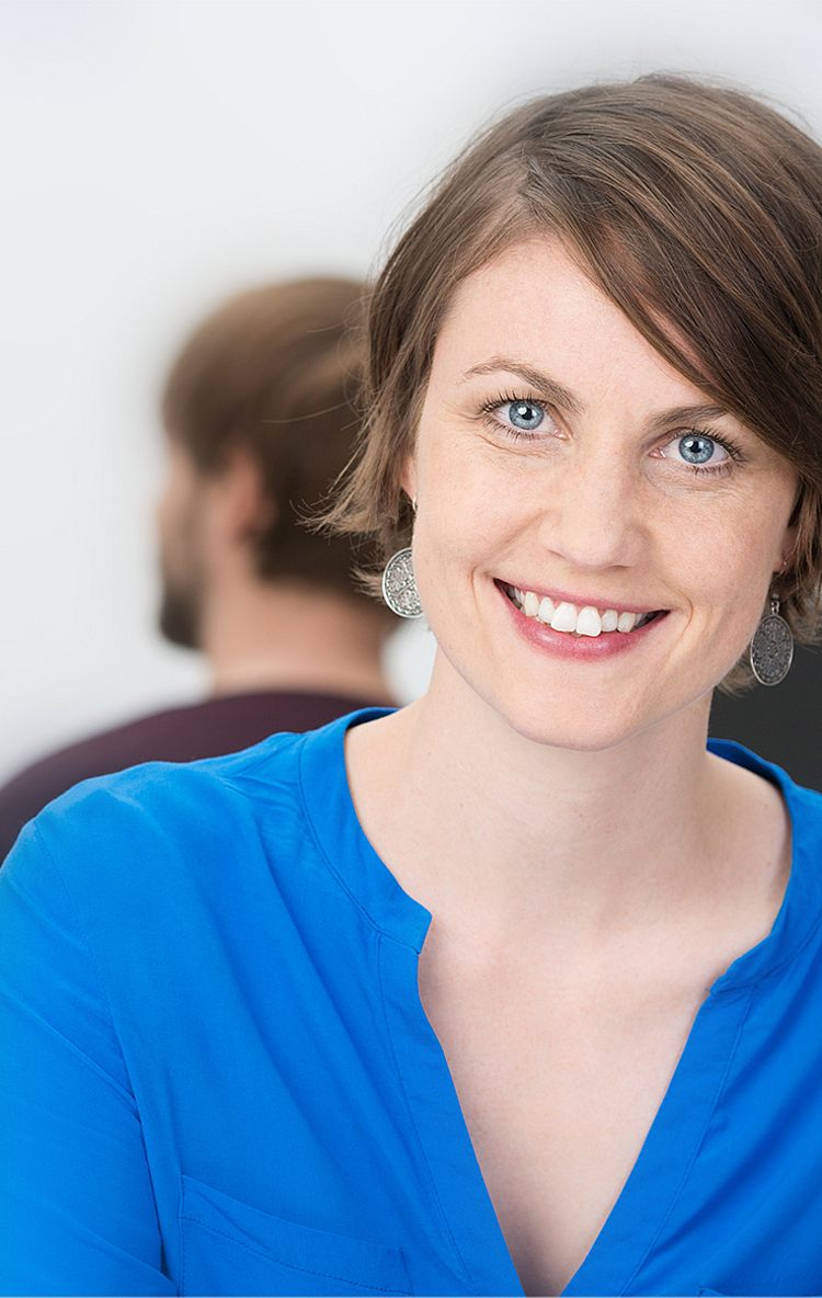 Eine lächelnde Frau mit kurzen, braunen Haaren trägt ein blaues Hemd. Sie sitzt in einem modernen Büro und schaut direkt in die Kamera. Im Hintergrund sind unscharf zwei weitere Personen bei der Arbeit zu sehen.